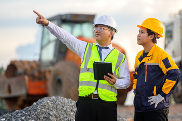 A civil engineer pointing direction to a worker on a construction site with heavy machinery, a road roller, and gravel, industrial project planning.