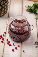 Glass teapot with hot berry tea and cranberries on wooden surface, next to mint and dried tea leaves