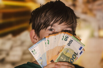 Smiling boy proudly holding a fan of euro banknotes in 20, 50, and 100 denominations, symbolizing wealth, savings, finance, and prosperity.