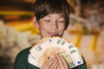 Smiling boy proudly holding a fan of euro banknotes in 20, 50, and 100 denominations, symbolizing wealth, savings, finance, and prosperity.