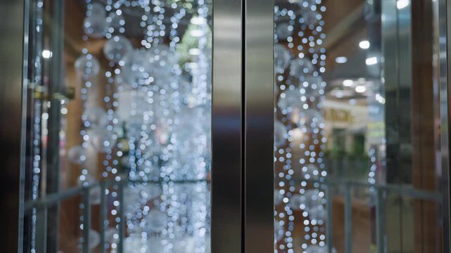 Close up of elevator door inside shopping mall with shiny metallic surface reflecting glowing ornaments creating bright holiday atmosphere with blurred decorative background