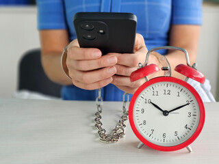 Holding a smartphone while chained to a clock in modern workspace offers a reflection on time...