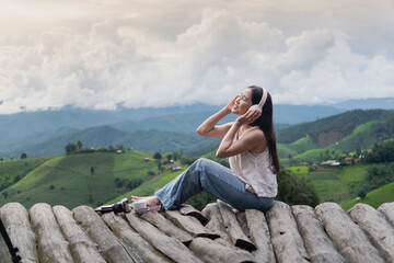 Naklejka premium A asian young woman sits on wooden logs in the mountains, wearing headphones, enjoying music and the scenic green valley view.
