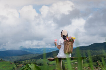 A woman in a white dress with a flower basket on her back, sitting by green hills under a cloudy sky