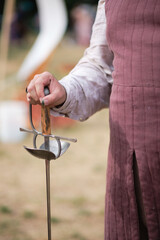 Medieval reenactor in period clothing holding a rapier with a wooden hilt and protective guard. Close-up of hand gripping sword during historical event outdoors on grass.
