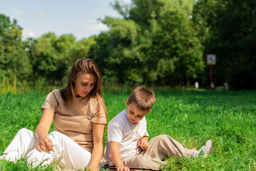 Fototapeta premium Mother helping her small son reading book about cars and transformers spending time on nature sitting on grass on summer day.