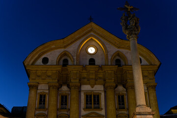 Monastery in Ljubljana at night. Uršulinski samostan by Kongresni trg. Illuminated clock on historic building in the city center. Detailed architecture with monument Pillar of the holy Trinity.