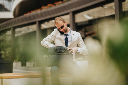 A business professional multitasks in an outdoor setting, handling a bag while conversing on the phone. The stylish modern architecture and greenery underline professionalism and dynamic city life.