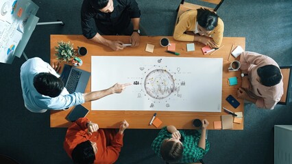 A diverse group of professionals gathers around a wooden table during a strategy planning session. They engage in brainstorming ideas and discussing various concepts with charts and notes. Raster
