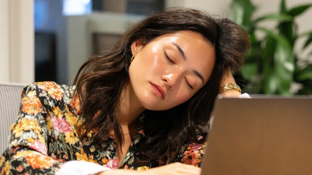 A woman with dark hair is resting her head on her hand at a desk. She appears tired, surrounded by a contemporary office backdrop and greenery. The warm lighting adds a calm atmosphere.