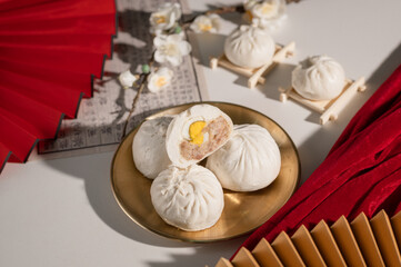 Steamed buns on a gold plate, with one cut open to reveal its filling. Surrounding decor includes red fans and delicate flowers, creating an elegant presentation.
