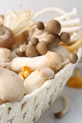 Different raw mushrooms in wicker basket on white tiled table, closeup