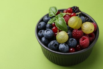 Different ripe juicy berries and basil in bowl on green background, closeup. Space for text