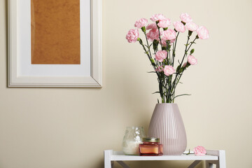 Beautiful carnation flowers in vase and candles on white stand near beige wall indoors