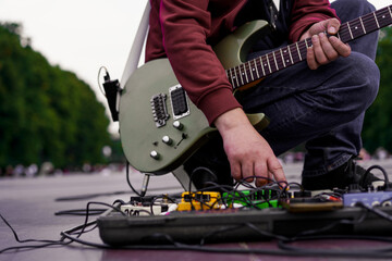 Musician plays electric guitar while adjusting effects pedals in a lively park setting during the evening hours