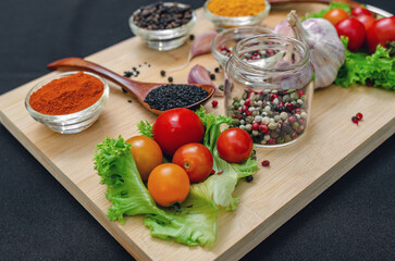 Cherry tomatoes, garlic, dry spices in bowls and spoons on  wooden cutting board on black backdrop.