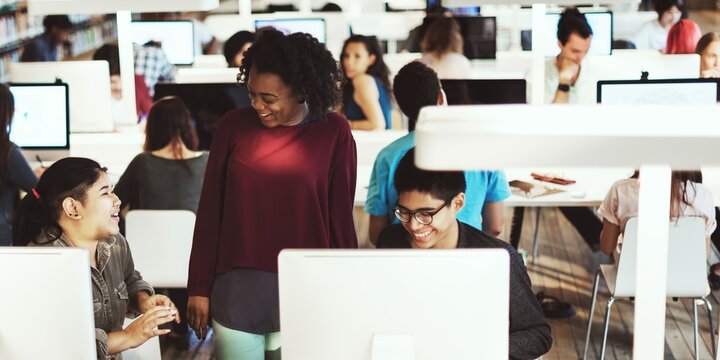 Teacher and diverse students studying in a library with computers. Diverse young adults studying and learning with computers. Students talk with teacher in library with computers. Learning in library