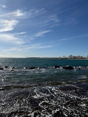 A scenic coastal view with turquoise waves crashing against rocks, a clear blue sky streaked with clouds, and a distant city skyline along the horizon.