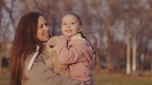 child teddy bear, kid mother, little girl, hug kid, carry in her arms, happy family, kid with mummies city park, favorite soft toy baby, mothering and taking care her daughter, mom laughs and smiles.