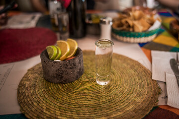 Frontal photo of a delicious crystalline mezcal in its glass caliber, accompanied by worm salt, oranges, and lemon served in a small molcajete.
