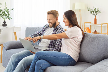 Happy husband and wife at home resting on sofa holdinng laptop to choose goods online, young family couple using pc, discussing food order, flat renovation, planning trip vacation, planning wedding
