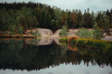 Obraz premium Serene Forest Lake Reflection. A tranquil lake reflects the lush green and brown trees of a surrounding forest under a slightly overcast sky. 