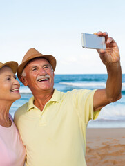 An elderly couple smiles brightly as they take a selfie on a beautiful beach. The sun shines down, and the ocean waves create a relaxing backdrop for their joyful moment.