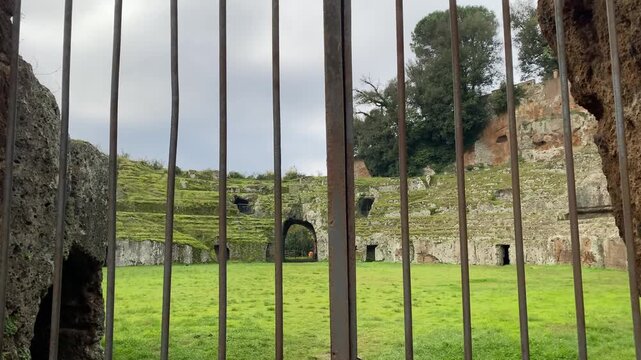 Camera approaching metal gate and showing old ruined arena stage for battles overgrown with greenery, abandoned amphitheatre being a part of medieval history, cultural monument seen through the