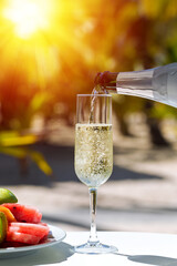 Pouring a champagne into flute glass, amazing fizz and bubbles in sparkling wine, table served with fruit plate at outdoor restaurant  on sunset with sun beams