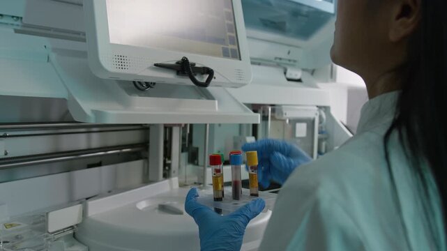 Female laboratory researcher holding blood test tubes and adjusting an automatic biochemistry analyzer machine, over the shoulder shot.