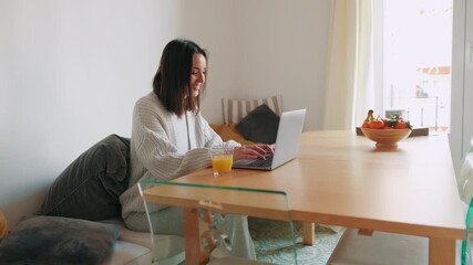 A young woman sits at her dining table, working remotely. She types on her laptop and enjoys a glass of orange juice. - Powered by Adobe