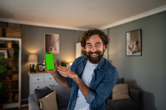 Smiling man presenting smartphone with green screen at home - Powered by Adobe