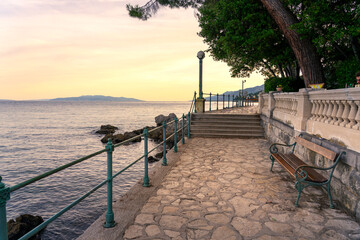 beautiful seaside promenade at sunset with stone pavement, benches, railing, and a calm sea. This is the Lungomare promenade in Opatija, Croatia