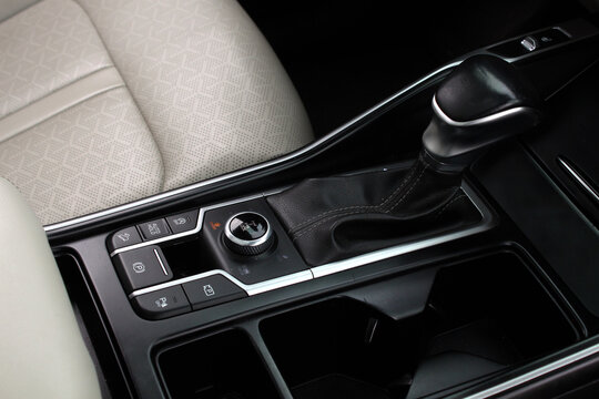 Black car center console with leather gear shifter, rotary drive mode selector, and control buttons, accented by silver trim and beige patterned seat upholstery.