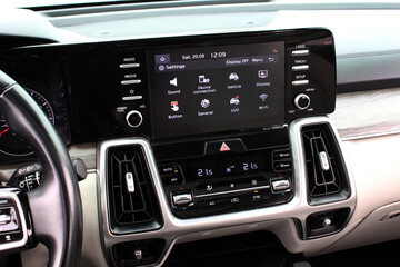 Car interior center console with touchscreen showing settings menu, flanked by physical media buttons and knobs, dual-zone climate control, and sleek silver-trimmed air vents. Screen multimedia system