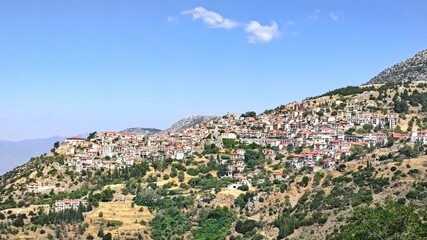 Obraz premium Panoramic view of Arahova, Greece, a picturesque mountain town on the slopes of Mount Parnassus, with red-tiled houses, stone buildings, and lush greenery under a clear blue sky.