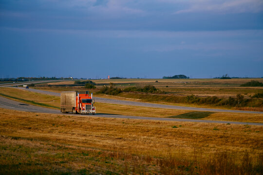 A large freight truck drives along a prairie highway in Saskatchewan, Canada, with wide open farmland and distant vehicles under dramatic evening light.