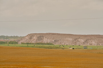 A large red potash tailings pile stretches across the horizon behind farmland with hay bales and a small building under cloudy skies in Saskatchewan, Canada.