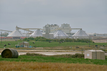 Industrial potash processing plant in Saskatchewan, Canada, with large domed storage structures, pipelines, and construction equipment in the foreground under cloudy skies.