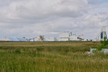 Main processing building of a potash plant in Saskatchewan, Canada, with white industrial architecture, ventilation stacks, and smoke rising under dramatic dark clouds.