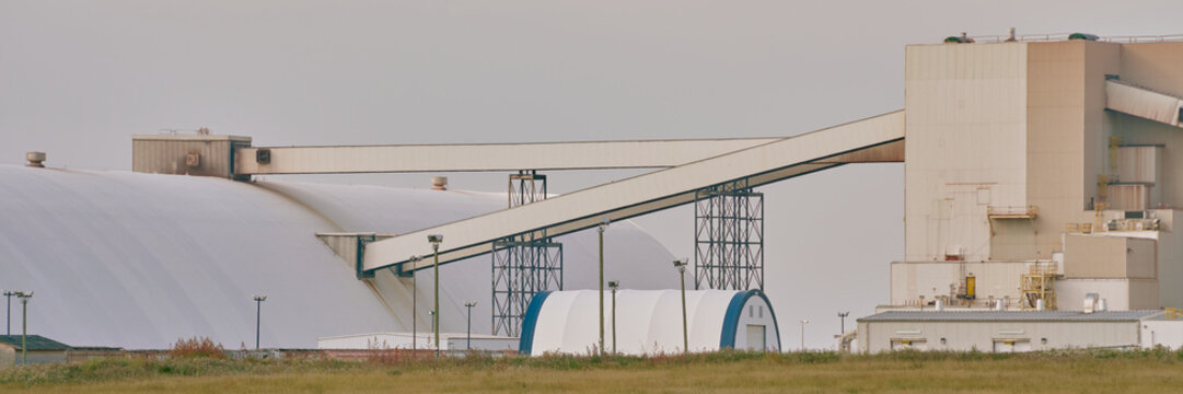 Main processing building of a potash plant in Saskatchewan, Canada, with white industrial architecture, ventilation stacks, and smoke rising under dramatic dark clouds.