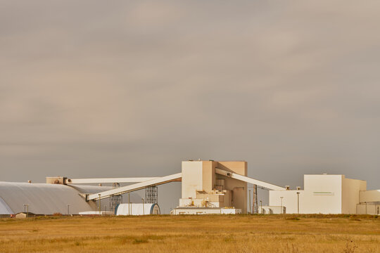 Modern industrial mining facility in Saskatchewan, Canada, featuring white factory buildings, conveyor systems, and a tall shaft tower against a cloudy sky with grassy foreground.