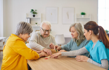 Happy smiling senior elderly people group sitting at table in retirement home collecting geometric figures with young friendly nurse. Dementia prevention and leisure time in nursing home concept.