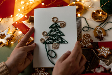 Handmade New Year paper postcard with embroidering of christmas fir tree. Holiday family time and gift for beloved. Gingerbread cookies, lights, wooden background. Zero waste, recyclable materials