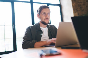 Fototapeta premium Modern male freelancer types on laptop in bright loft office, concentrating on startup tasks, surrounded by digital devices and creative documents, reflecting focused workflow.