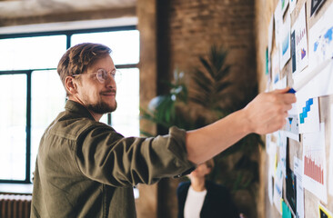 Smiling businessman points confidently at UX chart on idea wall, presenting design direction during tech project review in casual, collaborative digital workplace.