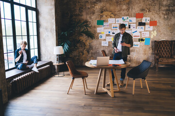 Young team deeply focused on digital concept, woman reviewing tablet notes while man checks smartphone near planning table covered in documents and tech devices.