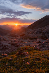 Sunset over rocky mountain landscape with glowing sun, dramatic clouds, and warm evening light on green moss foreground