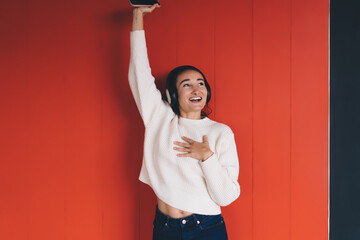 Young happy woman enjoying music in headphones, raising phone, smiling with joy against red wall,...