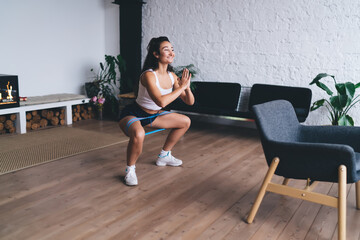 Focused young woman doing deep squat with resistance band at home, representing strength, self-discipline and mental balance through independent fitness practice.